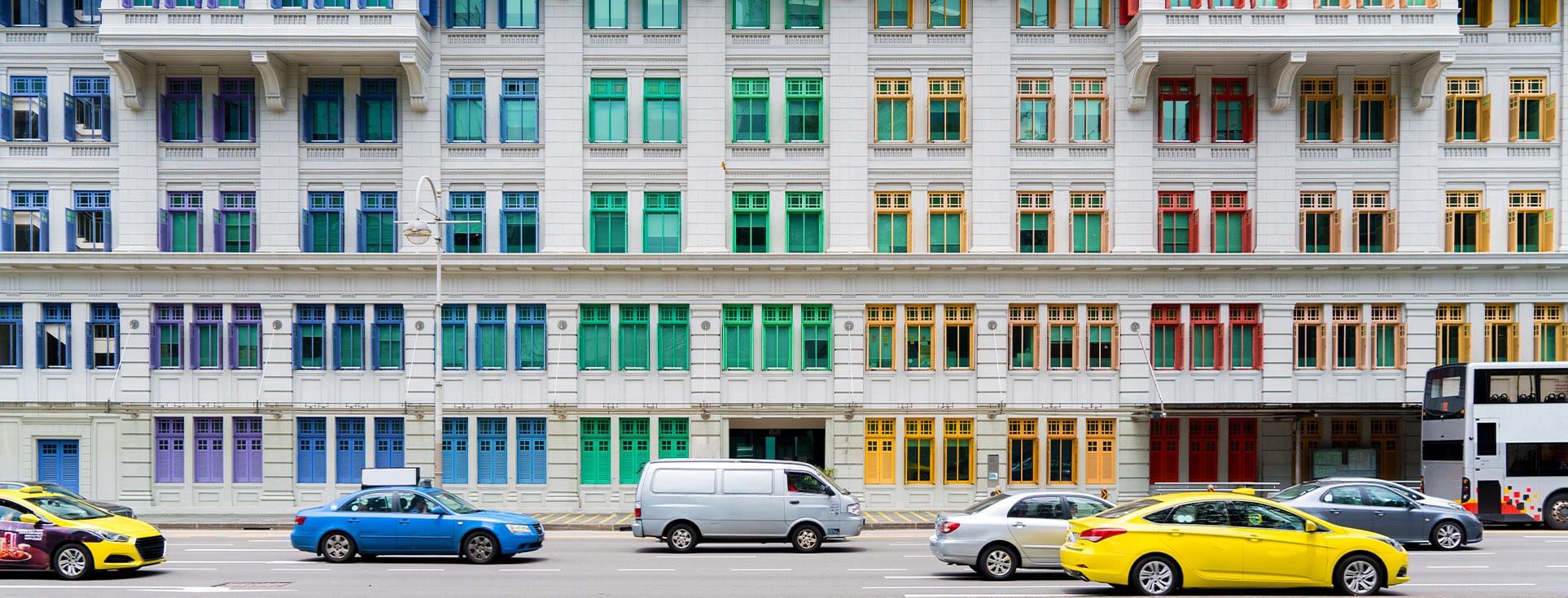 White building with colorful windows in purple, blue, green, yellow, and red, with cars and a bus on the street in front.